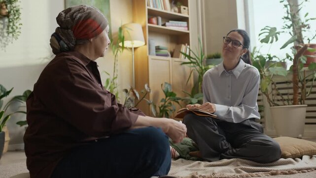 Full shot of mature female patient wearing headscarf sharing her feelings with middle-aged Caucasian female psychologist while sitting on floor in cozy green office
