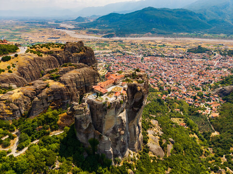 Aerial view of monasteries perched atop towering rock formations, contrasting with the sprawling town below and distant mountains, Meteores, Trikala, Greece.