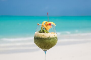 An exotic tropical coconut cocktail stands on the sun-drenched beach sand as a cold, refreshing summer drink against the backdrop of the blue sea and sky © Najmul
