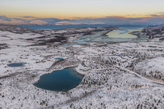 Aerial view of snow-dusted landscapes meeting icy waters under a soft, fading light, Lyngen, Troms, Norway.