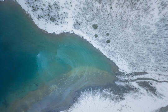 Aerial view of turquoise waters meet the snow-laden shores, a stark contrast of cool blues and icy whites in the winter landscape, Lyngen, Troms, Norway.