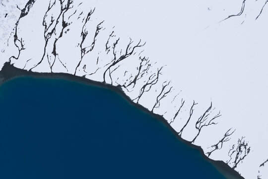 Aerial view of dark, skeletal lines of glacial streams carving through the stark white snowfields meeting the deep blue waters of a fjord, Lyngen, Troms, Norway.