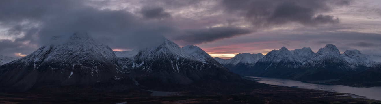 Aerial view of snow-dusted peaks pierce through a veil of twilight, casting long shadows across the still waters below, Lyngen, Troms, Norway.