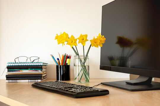 Modern office desk with computer monitor, keyboard, stack of notebooks, pencils and yellow daffodils. Stylish workplace scene with copy space, home office, productivity, remote work and study concept.