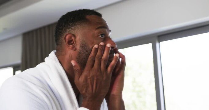 Mature African American man standing at window wearing white bathrobe, massaging under-eye skin
