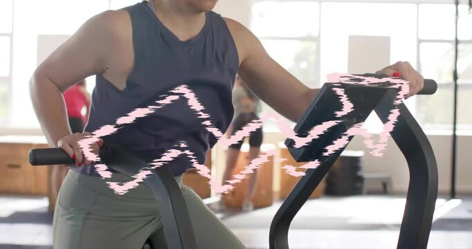 Cam panning up revealing woman gripping handles pedaling fan bike in gym, pink scribble flickering