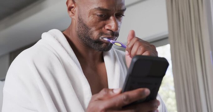 Man is checking black phone and brushing teeth with purple-yellow brush near window, smiling