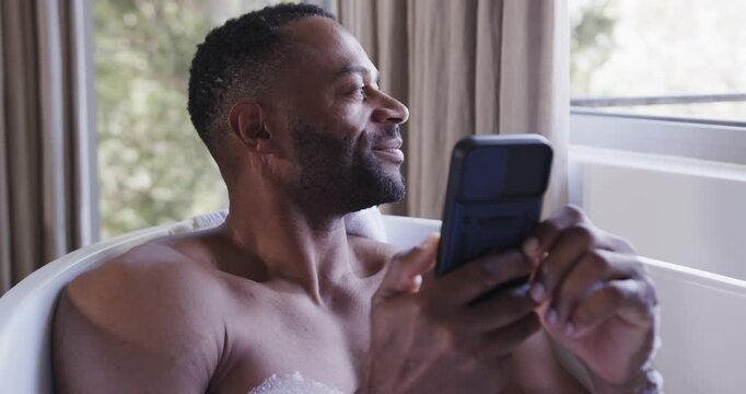 African American man sitting in tub shirtless, holding phone, scrolling beside window with bubbles