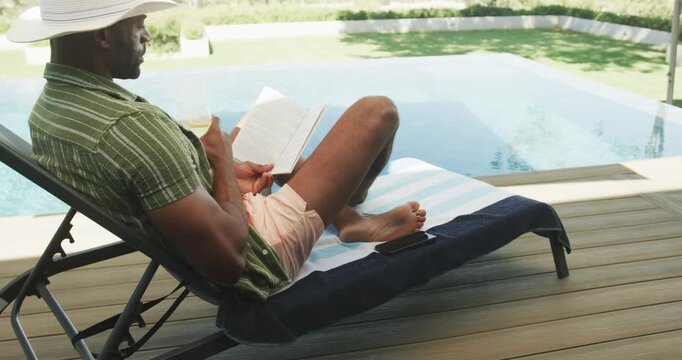 Reading mature man reclining at private poolside deck, wearing white hat, holding book and glass