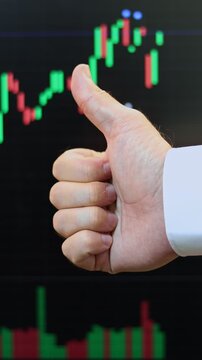Close up of a male hand showing thumb up gesture against a stock market terminal. Businessman making approval sign in front of trading screen with candlestick chart. Financial success and profit conce