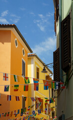 Obraz premium Vibrant colored houses in a narrow street in Piran, Slovenia, decorated with international flags of different countries. A beautiful blue sky completes this festive scene.