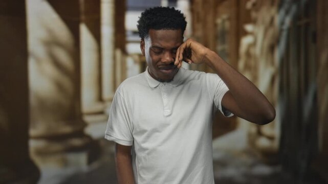African american man wipes tear from eye in a weathered stone building corridor framed by columns outdoors; sadness.
