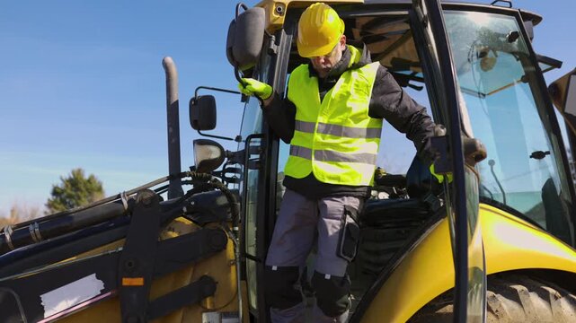 A construction worker in protective gear climbs down from a backhoe loader cabin during active work at an outdoor construction site.