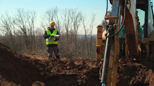 A construction engineer in safety gear stands beside a backhoe loader and reviews project documents during excavation work on a muddy construction site.