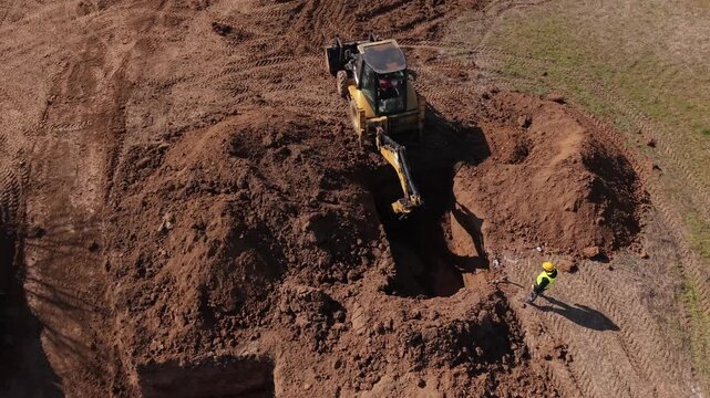 Aerial drone view of a construction engineer in safety gear supervising a backhoe loader near a large excavation pit on an active earthworks site.