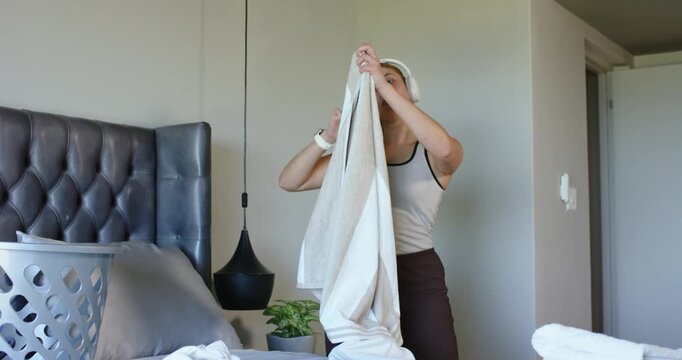 African American woman wearing headphones folding towels on bed, holding striped towel and stacking