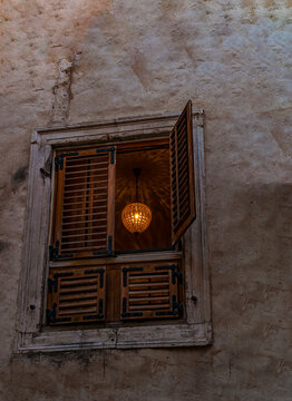 Croatia, Europe: crystal chandelier with the light on glimpsed from a wooden shuttered window in the alleys of the old town of Split. Stone wall, ancient building 
