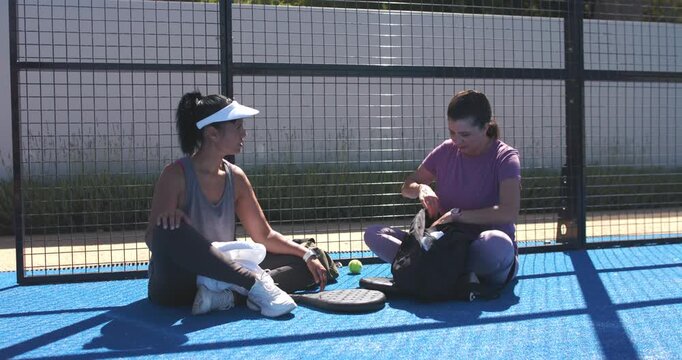 Diverse female pair sitting on blue court rummaging black duffel, packing paddles after match