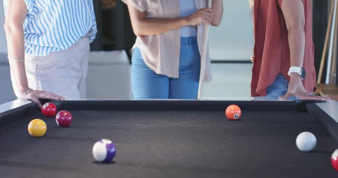 Camera pulling back, three women holding cues, leaning, eyeing balls at pool table, planning shots