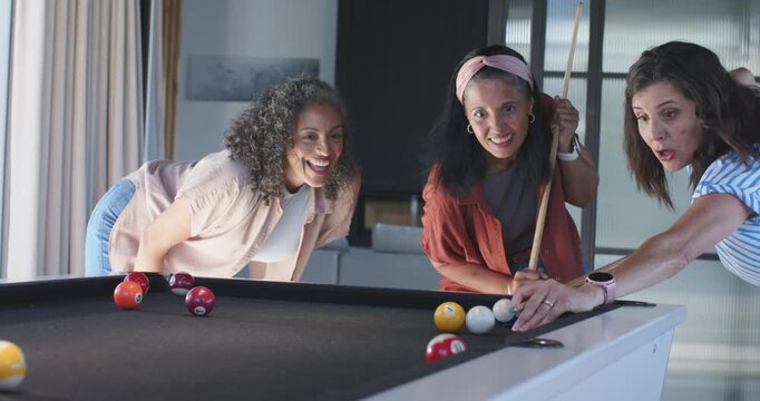 Receiving guidance, right woman in striped top hitting cue-ball on pool table at home for fun