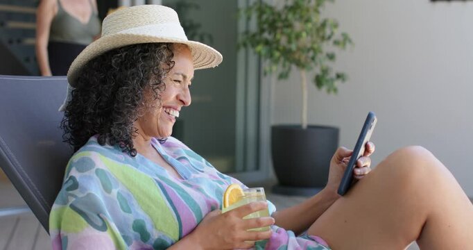 Seated woman receiving text, scrolling phone, laughing and sipping drink as companion walking by