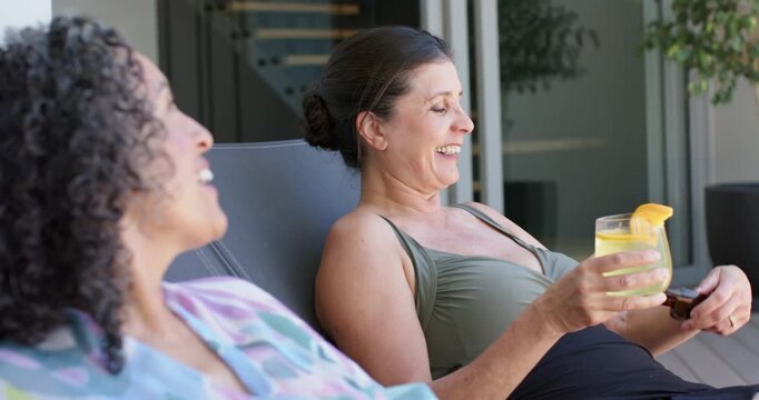Smiling right woman in green swimsuit raising glass with sunglasses, sharing laugh on home deck