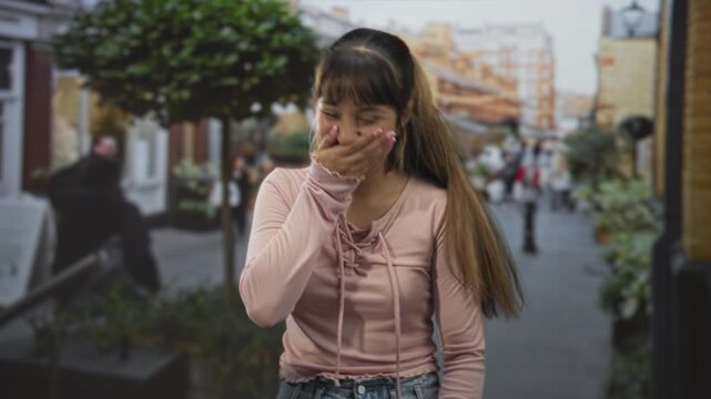 Young woman in pink top with hand covering mouth laughing on a busy street lined with storefronts and planters; shy amusement.