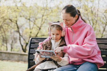 Happy mother and daughter sitting on a park bench watching tablet.