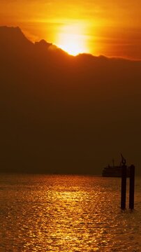 Vertical video. Golden Sunset Above Dark Clouds Over Calm Ocean With Passenger Ferry Silhouette on Horizon and Two Navigation Poles in Water. Warm Reflection on Sea Surface in Dramatic Evening Seascap