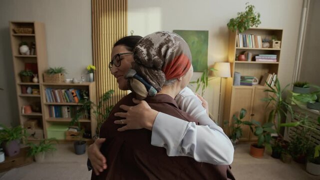 Medium slowmo shot of empathetic middle-aged Caucasian female psychologist hugging mature Asian female patient in headscarf, finishing successful therapy in modern office