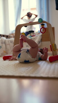 Baby lying on mat with legs raised, playing beneath wooden toy arch, engaged in early sensory and motor exploration, calm moment in cozy home environment