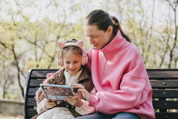 Happy mother and daughter sitting on a park bench watching tablet.