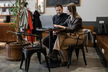 Fototapeta premium Professional man and woman discussing plans and using laptops on wooden bench in office with tree decor. Team discussion, planning, laptop, tree decor, workspace.