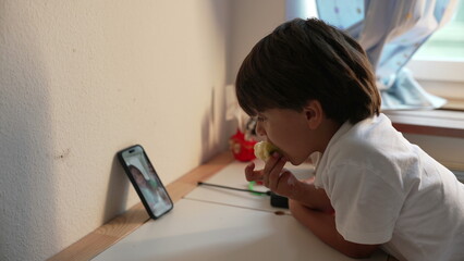 Naklejka premium Young boy eating apple while watching phone screen during video call, casual family moment, digital connection, childhood interaction in cozy home environment
