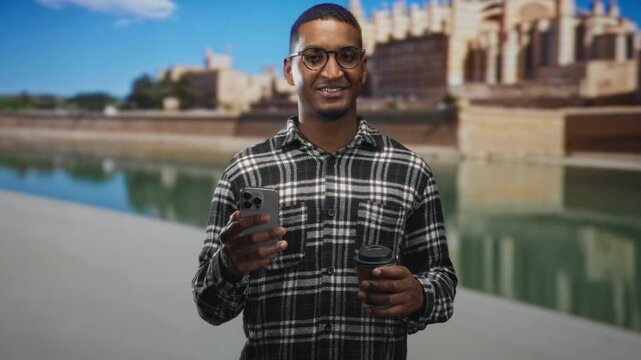 Man holding smartphone in right hand and takeaway coffee cup in left, smiling on a street by a historic building waterfront; contentment.