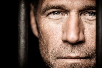 Fototapeta premium Intense close-up of a man behind bars, conveying a feeling of confinement, featuring detailed skin texture, piercing eyes, and a somber expression under dramatic lighting.