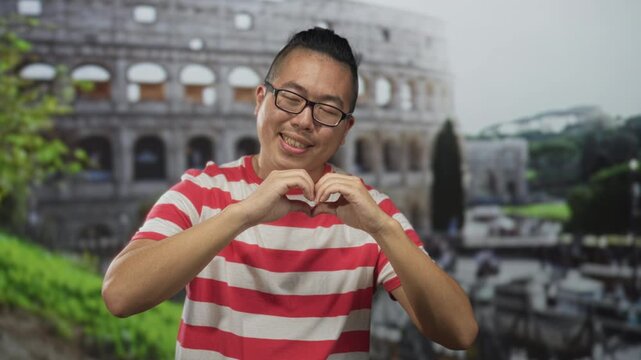 Man making heart with hands in front of colosseum building, wearing red striped t shirt and glasses while smiling with closed eyes; travel love.