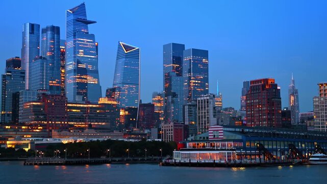 Panoramic view of illuminated Hudson Yards skyline at night. Wide angle of Manhattan west side skyscrapers with glowing windows and neon lights reflected on river surface.