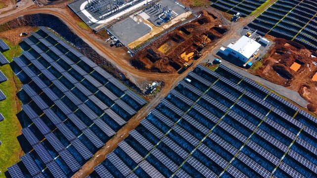Aerial view of solar power plant construction site and substation. High angle view of photovoltaic panels, electrical substation, excavators and dirt roads at industrial site.
