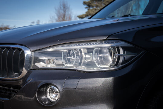 Close up of BMW X5 front left corner shows twin projector headlamps, kidney grille, fog light, and LED bars. Daylight reflects sky and bare trees in cool season.