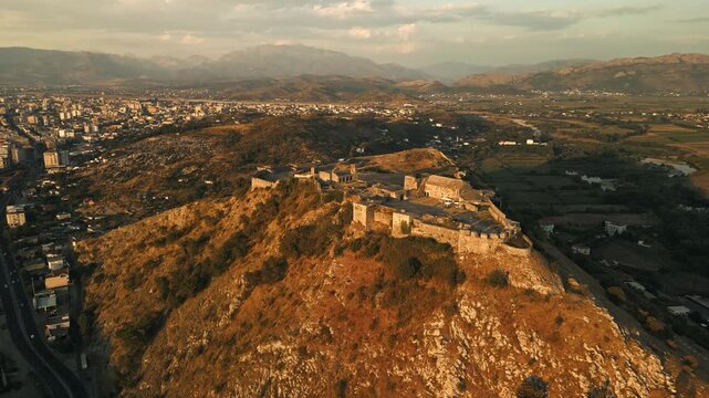 Aerial orbit of Rozafa Castle on hill overlooking Shkoder city and Albanian Alps at golden hour, Albania