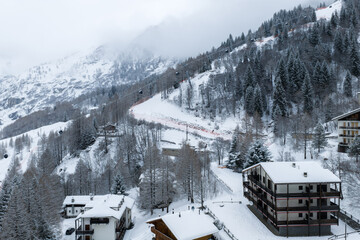 Aerial view of San Domenico in the Italian Alps shows lodges, a luxury loft hotel, gondola lift, orange fencing, steep pistes, dense spruce and larch, and winter activity. © Aerial Film Studio