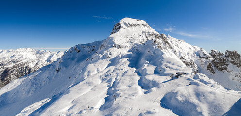 Fototapeta premium Aerial view shows San Domenico ski resort in the Italian Alps. Groomed pistes line a snow peak, ski lifts and a small station sit on a ridge, skiers descend broad slopes.