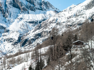 Aerial view shows San Domenico ski resort in the Italian Alps, rugged snow cliffs, larch dotted slopes, gondola lifts in motion, skiers' tracks, and a chalet hotel. © Aerial Film Studio