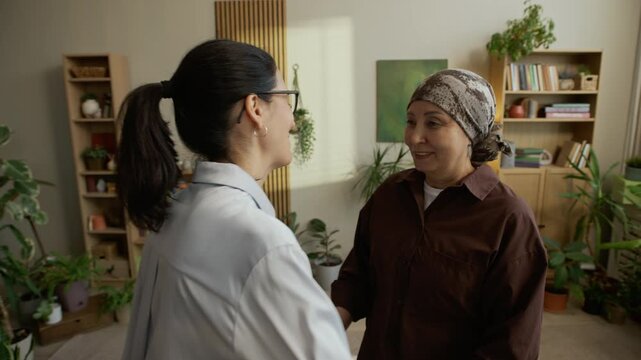 Medium slowmo shot of grateful mature Asian female patient in headscarf hugging middle-aged Caucasian female psychologist after finishing successful therapy in cozy green office