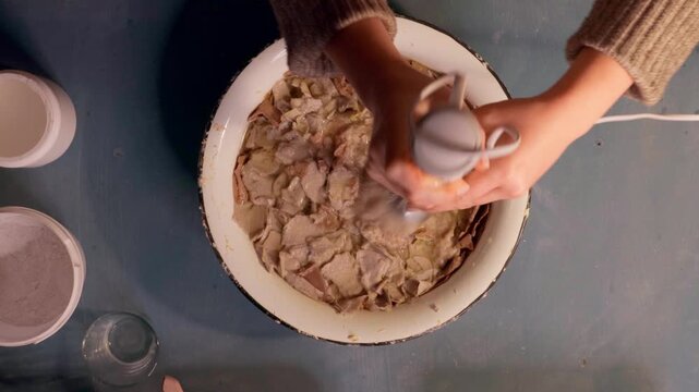 Step 2 of papier mache making process. Hands soaking torn egg carton pieces in water to soften recycled paper for craft pulp mixture. DIY recycled paper craft preparation