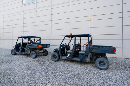 Industrial side by side utility carts parked near a modern exterior wall on crushed stone ground, showing cargo beds and safety flags in place