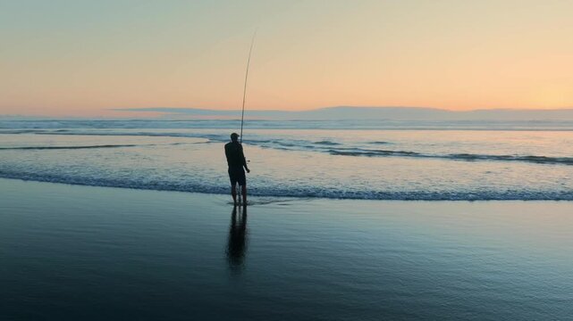 A lone fisherman casts his line into the surf at sunset in Piha, Auckland, New Zealand. He is hoping to catch fish for dinner, enjoying the peace of the ocean.