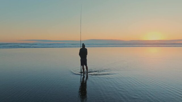 A lone fisherman casts his line into the surf at sunset in Piha, Auckland, New Zealand. He is hoping to catch fish for dinner, enjoying the peace of the ocean.