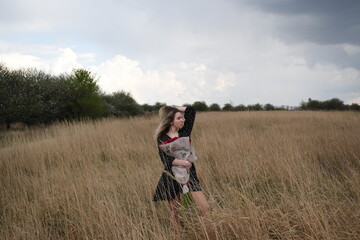 Young woman in black polka dot dress holding bouquet of red roses in autumn field under dramatic stormy sky.